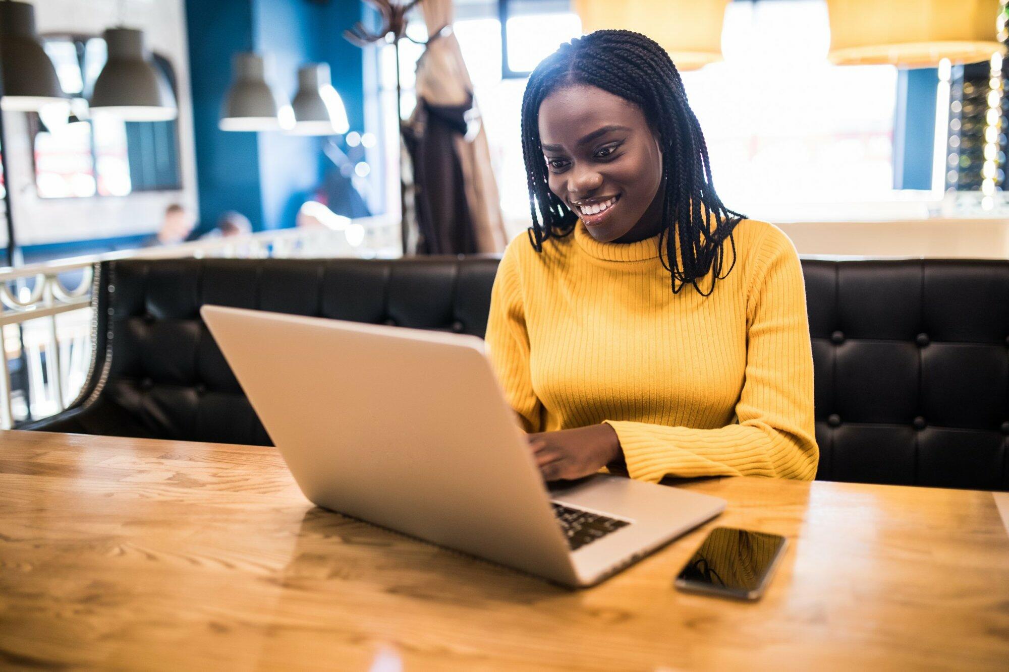Human and technology. Young african woman typing on laptop, texting friends via social networks. Student girl browsing Internet, using free wi-fi, sitting at cafe near window on sunny day